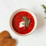 Top-down view of traditional borscht soup served with dill and slices of brown rye bread on a white surface.