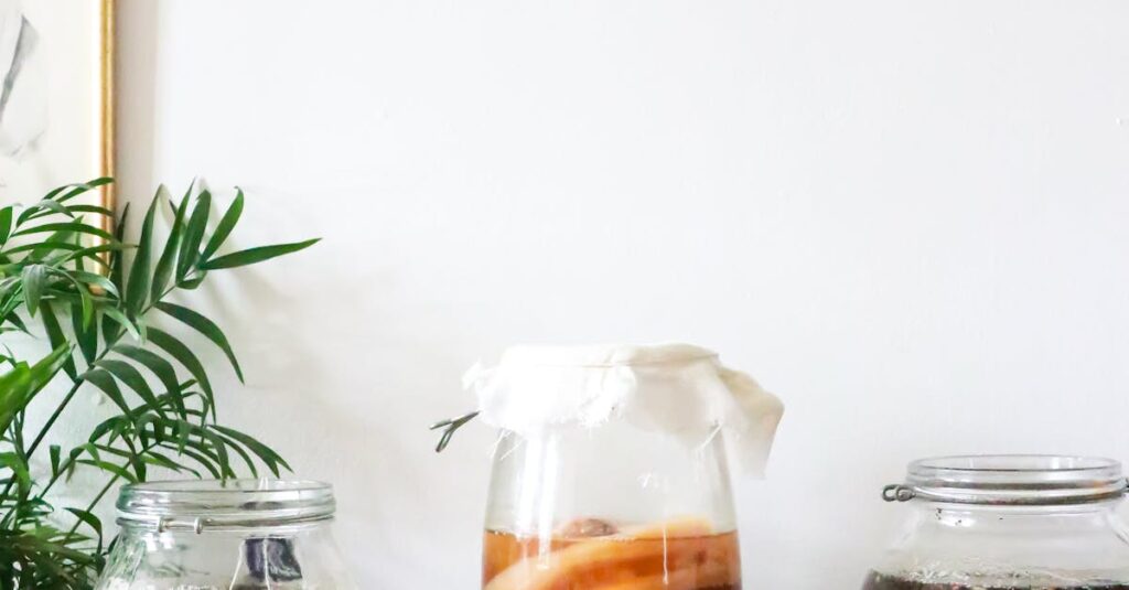 Glass jars with infused liqueurs with ingredients placed on wooden table near green plant near wall with decorations at home